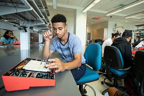 Student is sitting in a lab with other people with a circuit board in front of him. 