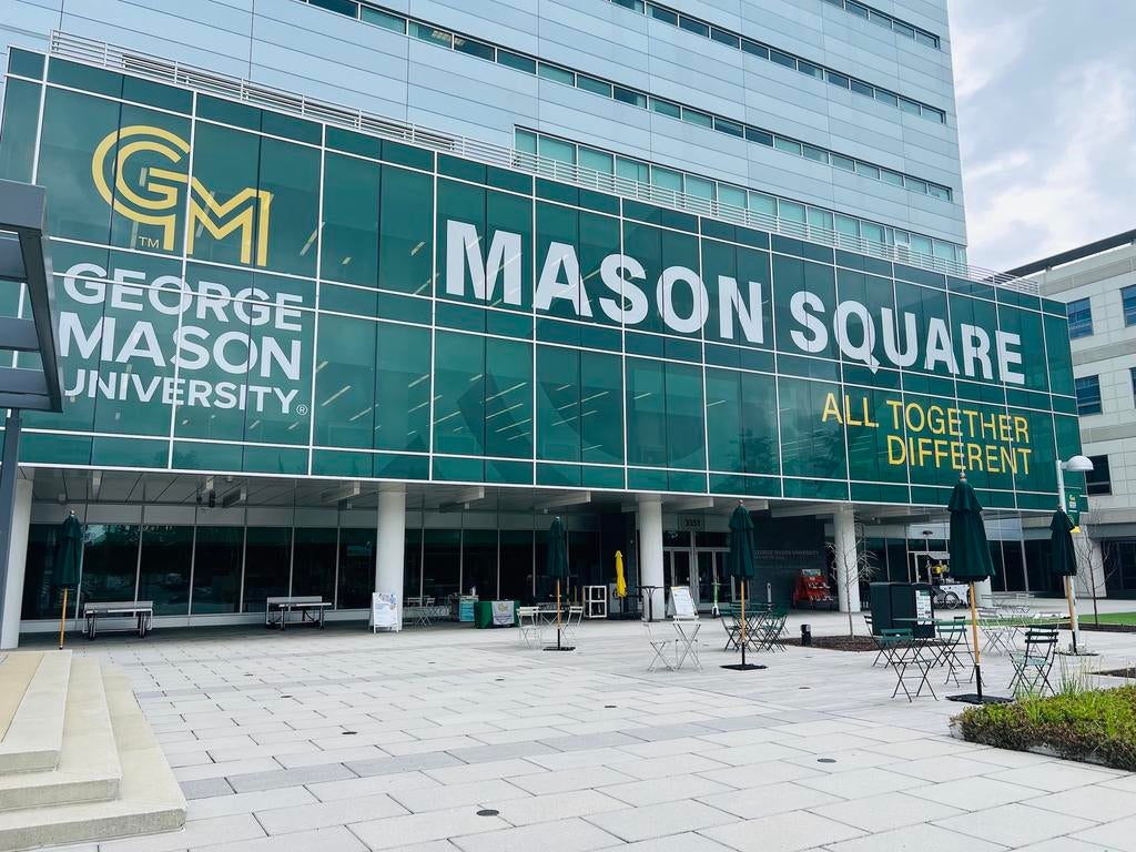 Exterior view of a modern George Mason University building at Mason Square, featuring large green-and-white signage with the GMU logo, the words “Mason Square,” and the slogan “All Together Different.” The foreground includes an open plaza with tables, chairs, and umbrellas.
